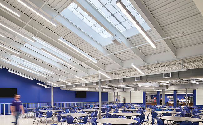MX4 continuous fixtures suspended at an angle below an open ceiling in a school cafeteria with a blue accent wall and round tables with blue chairs.